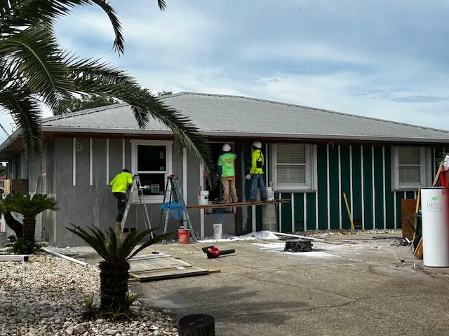 Workers renovating a green house exterior under a cloudy sky.