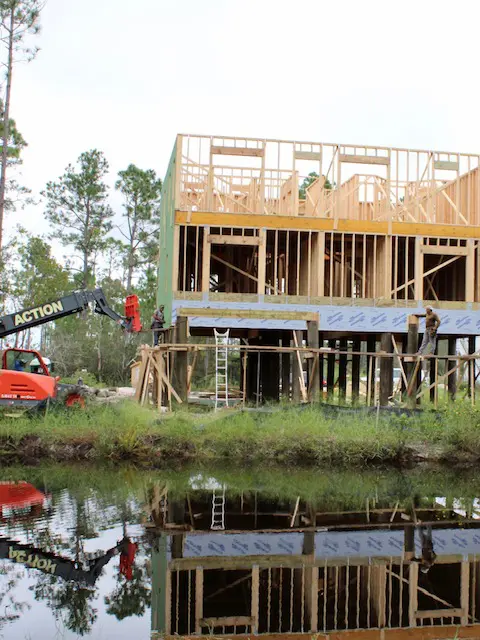 Wooden house frame under construction near a pond with trees in the background.